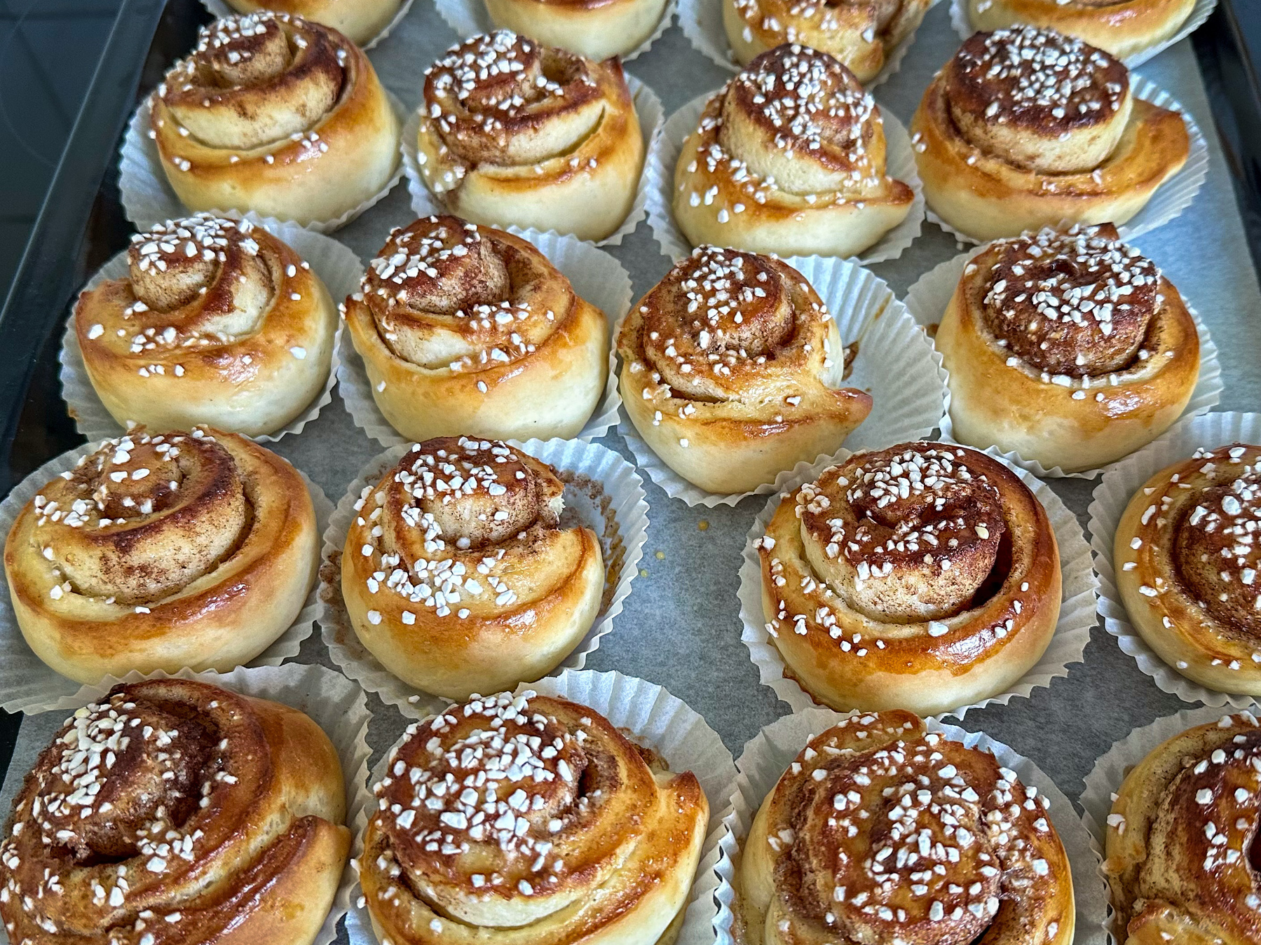 Auto-generated description: A tray of freshly baked cinnamon rolls topped with pearl sugar is displayed in baking cups.